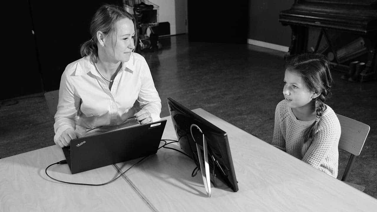 Et bilde i svart-hvitt av en kvinnelig lærer og en elev som sitter foran hver sin PC-skjerm og utfører en kartlegging